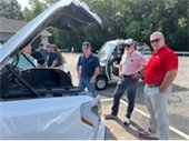 Several men stand in front of an electric F-150 open hood. An electric club car vehicle with a lift is in the back ground along with other people