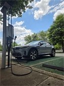 Shot from close to the ground, a grey Honda Prologue charges at the ACC public EV charger at Park and Ride. There are trees and a blue sky with white clouds.