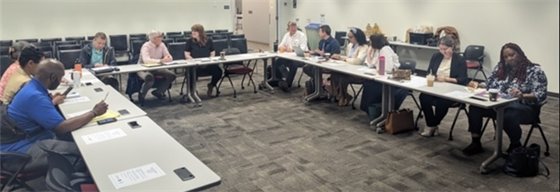 Several people sit in a half circle of chairs and tables chatting